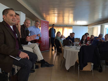A group of people sit around a table or stand and talk at the Tallberg workshop